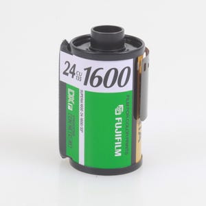 Puede incluir: Un rollo de película en color Fujifilm Superia 1600 para cámaras de 35 mm. El cartucho de película es negro y dorado con una etiqueta verde. La etiqueta muestra el texto "24 1600" y "FILM FOR COLOR PRINTS".