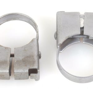 Könnte beinhalten: Zwei silberfarbene Metallrohrschellen mit einem kreisförmigen oberen Teil und einem rechteckigen unteren Gehäuse. Jede Schelle verfügt über einen horizontalen Gewindeschraubenmechanismus zum Festziehen der Öffnung mit matter Oberfläche.