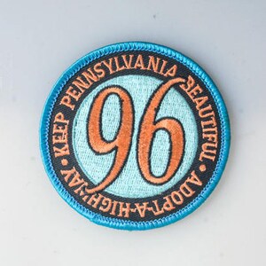 Könnte beinhalten: Ein runder blauer und schwarzer Aufnäher mit weißem Text, der "Adopt-A-Highway Keep Pennsylvania Beautiful" lautet, und einer großen orangen Zahl "96" in der Mitte.
