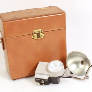 May include: A brown leather case with a brass latch, a silver flash unit with a white reflector, and a smaller silver flash unit with a white reflector.