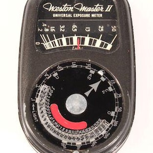 Puede incluir: Un exposímetro Weston Master II Universal Exposure Meter de color negro con una escala de luz analógica superior con marcas numéricas blancas y un dial circular inferior con un indicador rojo, números blancos y un botón central plateado.