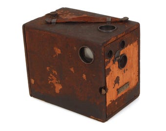 May include: A vintage box camera with a weathered, brown exterior. The camera has a rectangular shape with a lens, viewfinder, and a leather handle. The wood has a distressed appearance, suggesting age and use.