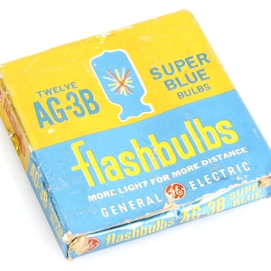 Op de afbeelding: Een vintage doosje met General Electric Super Blue flitslampen. De geel en blauwe doos draagt de tekst "flashbulbs" en "AG-3B". De doos is licht versleten, wat duidt op de leeftijd.