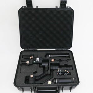May include: A black hard-shell carrying case with a foam-lined interior containing a black camera gimbal stabilizer, a tripod base, a handle, and various small components including a grey cable, all organized in custom-cut foam compartments.