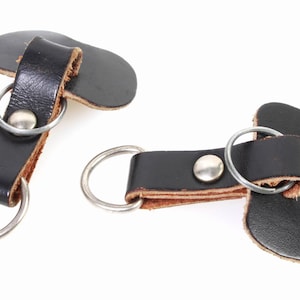 以下が含まれることがあります： 銀色の金属製のリングが付いた黒い革製のキーホルダーが2つ。各キーホルダーの上部には、小さな平らな革のループが付いています。