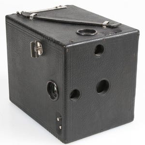 May include: A vintage black box camera with a textured surface. The camera has a rectangular shape with a leather strap and metal accents. It features several circular openings and a hinged latch.