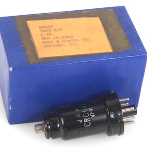 May include: A vintage electronic tube, labelled JAN CRC-6J VT-91, beside a blue box with a label. The box has text that reads: TUBE 6J7, 1 EA, and BELL & HOWELL CO. The tube is black with multiple pins.
