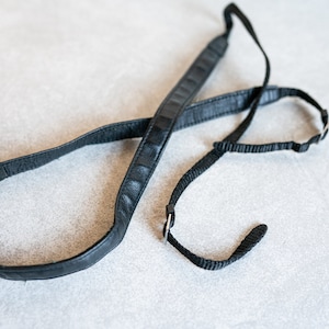 Puede incluir: Una correa de cámara de cuero negro con una correa de nylon negro adjunta. La correa está diseñada para usarse alrededor del cuello.