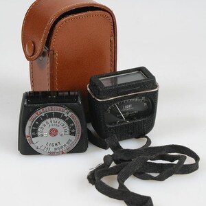 May include: A vintage General Electric light meter set featuring a black exposure calculator dial with f-stop markings, a black light meter unit with a needle display, and a brown leather carrying case with a snap closure and black fabric strap.