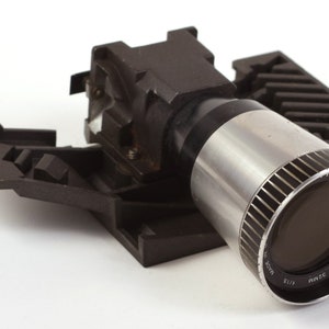 Puede incluir: Un objetivo de proyector negro y plateado con el texto "Bell & Howell" y "WIDE 0.5" impreso en el lateral.