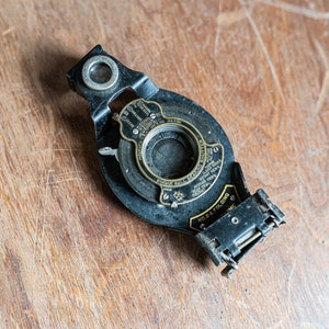 Puede incluir: Un obturador de cámara vintage negro con una abertura redonda y un dial de latón marcado con diferentes velocidades de obturación. El obturador está etiquetado como "No. R Folding" y tiene una pequeña ventana de visualización.