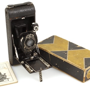 Op de afbeelding: Vintage zwarte Kodak camera met open balg, lens en zilverkleurig Kodak logo. Daarnaast een zwart-gouden doos en een boekje met de tekst "No. 1 and 1A Pocket Kodaks".