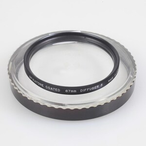 Puede incluir: Un filtro de lente de cámara negro Spiratone Coated 67mm Diffuser 5, dentro de una caja de plástico transparente con un borde marrón oscuro. El filtro tiene texto blanco y está diseñado para fotografía.