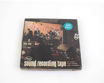 Könnte beinhalten: Vintage Tuck Tonbandkassette. Die Box zeigt ein Schwarz-Weiß-Bild eines Tonstudios mit arbeitenden Personen. Der Text auf der Box lautet "sound recording tape" und "600 feet". Die Box befindet sich auf weißem Hintergrund.