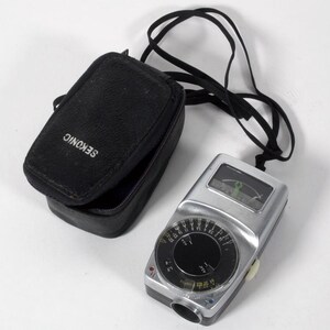 May include: A silver Sekonic light meter with a black circular dial featuring white numerical markings and a needle display, accompanied by a black zippered carrying case with a matching black neck strap attached to the device.