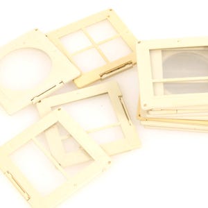 May include: A collection of vintage slide mounts in various designs. The off-white plastic frames have clear windows for viewing slides. Some have a circular opening, while others have a grid pattern. A stack of frames is visible.