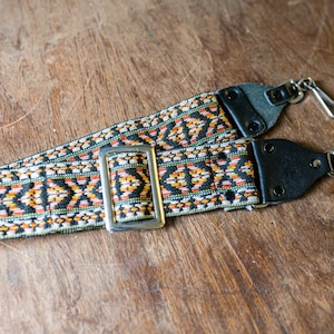 Op de afbeelding: Een zwart en oranje gepatenteerde cameraband met een zilveren gesp en twee metalen clips.