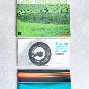 May include: Three Canon camera instruction manuals. The top manual is titled "Lens Wonderland" and features a green field with people walking. The middle manual is titled "Canon FD" and features a close-up of a camera lens. The bottom manual is titled "Canon Products Guide" and features a blue and orange design.