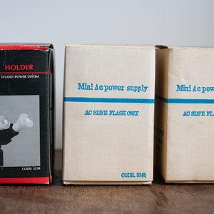 Puede incluir: Tres cajas que contienen equipo fotográfico. La caja de la izquierda es negra con texto blanco que dice "Twin Holder Studio Power System" y una imagen de un soporte de flash doble. Las dos cajas de la derecha son blancas con texto azul que dice "Mini Ac power supply AC Slave Flash Only" y el código "3348".
