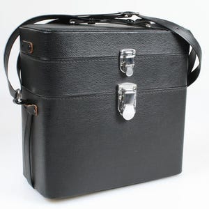 May include: A black leather camera case with a shoulder strap. The case has a rectangular shape with two silver metal clasps on the front. The leather has a textured appearance, and the strap is also black.