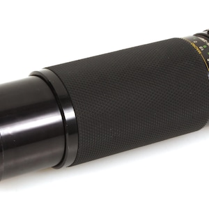 Puede incluir: Un objetivo de cámara Soligor negro con un agarre texturizado. El objetivo tiene un anillo frontal plateado con el nombre de la marca y el número de modelo. El objetivo tiene un anillo de enfoque con marcas de distancia.