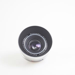 Pu&ograve; includere: Primo piano di un obiettivo fotografico con la scritta "Schneider-Kreuznach" e "Retina-Curtagon f/4 28mm". L'obiettivo ha un design nero e argento, su sfondo bianco.