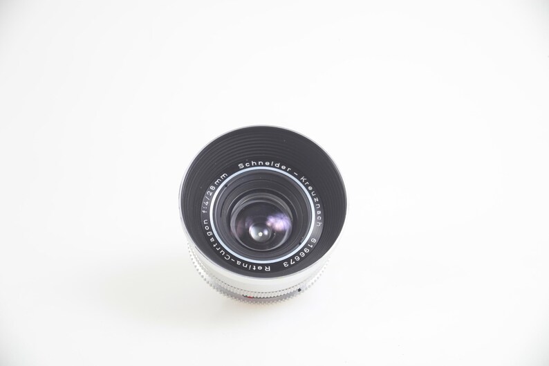 Pu&ograve; includere: Primo piano di un obiettivo fotografico con la scritta "Schneider-Kreuznach" e "Retina-Curtagon f/4 28mm". L'obiettivo ha un design nero e argento, su sfondo bianco.