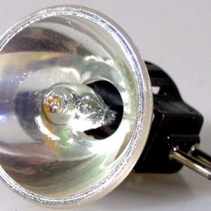 Puede incluir: Una bombilla halógena con un reflector circular de tono plateado y reflectante. El centro contiene una pequeña cápsula de vidrio transparente con un filamento visible. Una base negra se extiende desde la parte posterior, equipada con dos pines metálicos.