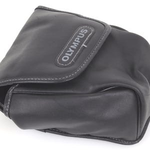 May include: Black leather camera case with the brand name "OLYMPUS" in white lettering. The case has a flap closure and white stitching around the edges. The case is designed to protect a camera.
