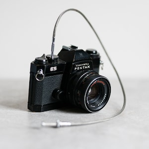 May include: Black Honeywell Pentax ES camera with a silver cable release. The camera has a lens with red and white lettering. The camera is on a light grey surface.