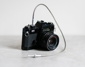 以下が含まれることがあります： 黒いHoneywell Pentax ESカメラとシルバーのケーブルレリーズ。カメラには赤と白の文字が入ったレンズが付いています。カメラは明るい灰色の表面にあります。