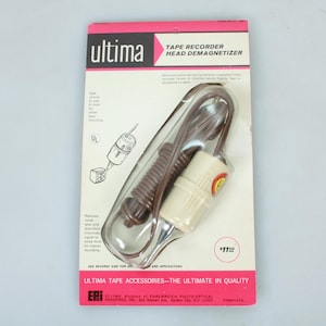 Puede incluir: Un desimantador de cabezal de grabadora de cinta blanco y marrón en un paquete de plástico. El paquete tiene el texto "ultima" y "TAPE RECORDER HEAD DEMAGNETIZER" en él. El paquete también tiene el texto "#11" en él.