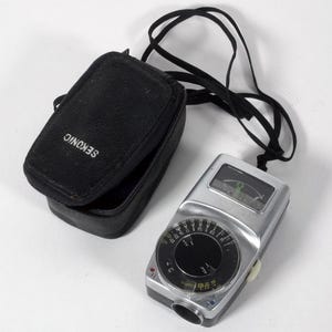 Puede incluir: Un fotómetro Sekonic vintage con una funda negra y una correa para el cuello. El fotómetro tiene un cuerpo plateado con un dial circular y una pequeña ventana que muestra las lecturas. La funda está abierta.