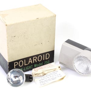 May include: A white box with the word "POLAROID" and the text "MK-LIGHT Model 250" printed on it. A white and gray flash unit with a black top is next to the box. A small, round flash bulb is in front of the box.