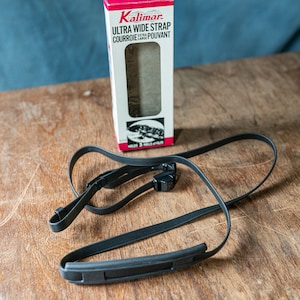 以下が含まれることがあります： 幅広のパッド付きセクションと2つの調整可能なクリップが付いた黒いカメラストラップ。ストラップは、「Kalimar Ultra Wide Strap Courroie Large Pouvant」というテキストが書かれた白い箱に梱包されています。