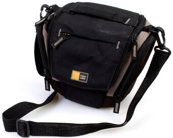 May include: A black and grey camera bag with a shoulder strap. The bag has a zipped pocket on the front with the text "CASO LOGIC" in yellow.