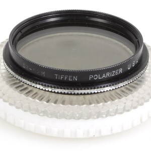 May include: A Tiffen Polarizer USA camera lens filter. The filter has a black ring with the text "Tiffen Polarizer USA" and a silver adjustment ring. It sits on a clear, textured base.