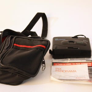 Op de afbeelding: Een zwarte camera, een zwarte schoudertas en een brochure. Op de camera staat het woord "PANORAMA". De schoudertas heeft een rode streep aan de bovenkant. De brochure zit in een plastic hoes.