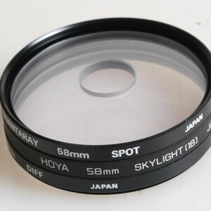 Puede incluir: Un filtro de lente de cámara circular negro con un centro de vidrio transparente que presenta un pequeño punto circular. El borde metálico tiene impreso el texto Hoya 58mm Skylight 1B, Japan y Diff. El filtro tiene un borde texturizado de 58mm.
