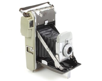 Könnte beinhalten: Eine Vintage-Polaroid-Kamera mit einem grauen und silbernen Gehäuse. Die Kamera ist geöffnet und zeigt den Balgen und das Objektiv. Die Kamera hat ein schwarzes Lederband.