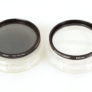 Może przedstawiać: Dwa filtry do obiektywów aparatu o średnicy 52 mm w plastikowych pudełkach. Filtr po lewej stronie to szary filtr polaryzacyjny. Filtr po prawej stronie to filtr przezroczysty.