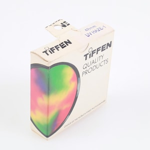 Op de afbeelding: Een Tiffen UV Haze-1 filterdoos. De doos is gebroken wit met het Tiffen-logo en "QUALITY PRODUCTS" aan de zijkant. Een hartvormig ontwerp op de voorkant heeft een kleurrijk, wazig patroon van roze, geel, groen en paars.