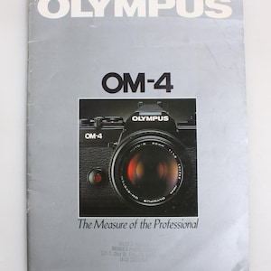 May include: A vintage Olympus OM-4 camera brochure. The cover is silver with the word "OLYMPUS" at the top. The text "OM-4" is prominently displayed, with a photo of the camera. The tagline "The Measure of the Professional" is below the camera.