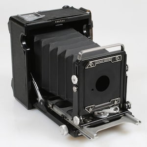 Peut inclure: Un appareil photo Century Graphic noir avec un soufflet. L'appareil photo a une poignée argentée et plusieurs cadrans et boutons. Le mot "GRAFLEX" est imprimé sur le dessus de l'appareil photo.