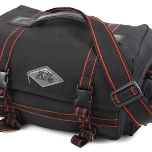 Könnte beinhalten: Schwarze Messenger-Tasche mit roten Akzentstreifen und -riemen. Sie verfügt über einen oberen Griff, einen verstellbaren Schultergurt und mehrere Schnallenverschlüsse. Die Tasche hat eine rechteckige Form mit einem Klappenverschluss und einem silbernen Logo-Patch "VINTAGE CLASSIC".