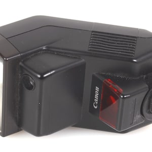 May include: Black Canon Speedlite flash unit. The flash has a rectangular head with a clear lens, a black body, and a red Canon logo. The flash has a hot shoe mount and a small red light.