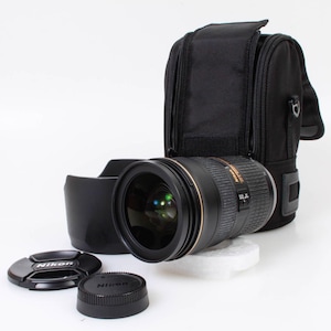 Op de afbeelding: Een zwart en goudkleurig cameralens met een zonnekap, lensdop en een zwarte draagtas. De lens ligt op een wit oppervlak. Op de lensdop staat het woord "Nikon".