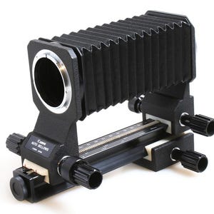 May include: A black Canon Auto Bellows camera accessory. The bellows has a ribbed design and is connected to a black base with adjustment knobs and a measurement scale. The Canon logo is visible.