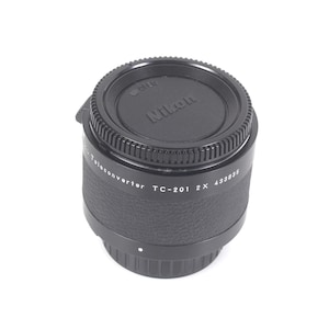 May include: A black Nikon TC-201 2X teleconverter lens accessory with a textured grip ring and a circular lens cap featuring the Nikon logo. The side of the cylindrical device displays the white text Nikon Teleconverter TC-201 2X 433835.