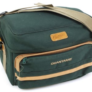 May include: A dark green Quantaray camera bag with a tan shoulder strap and accents. The bag has a rectangular shape with a front pocket and the word "Quantaray" printed on the front. The bag also has the word "Outgear" on a leather patch.